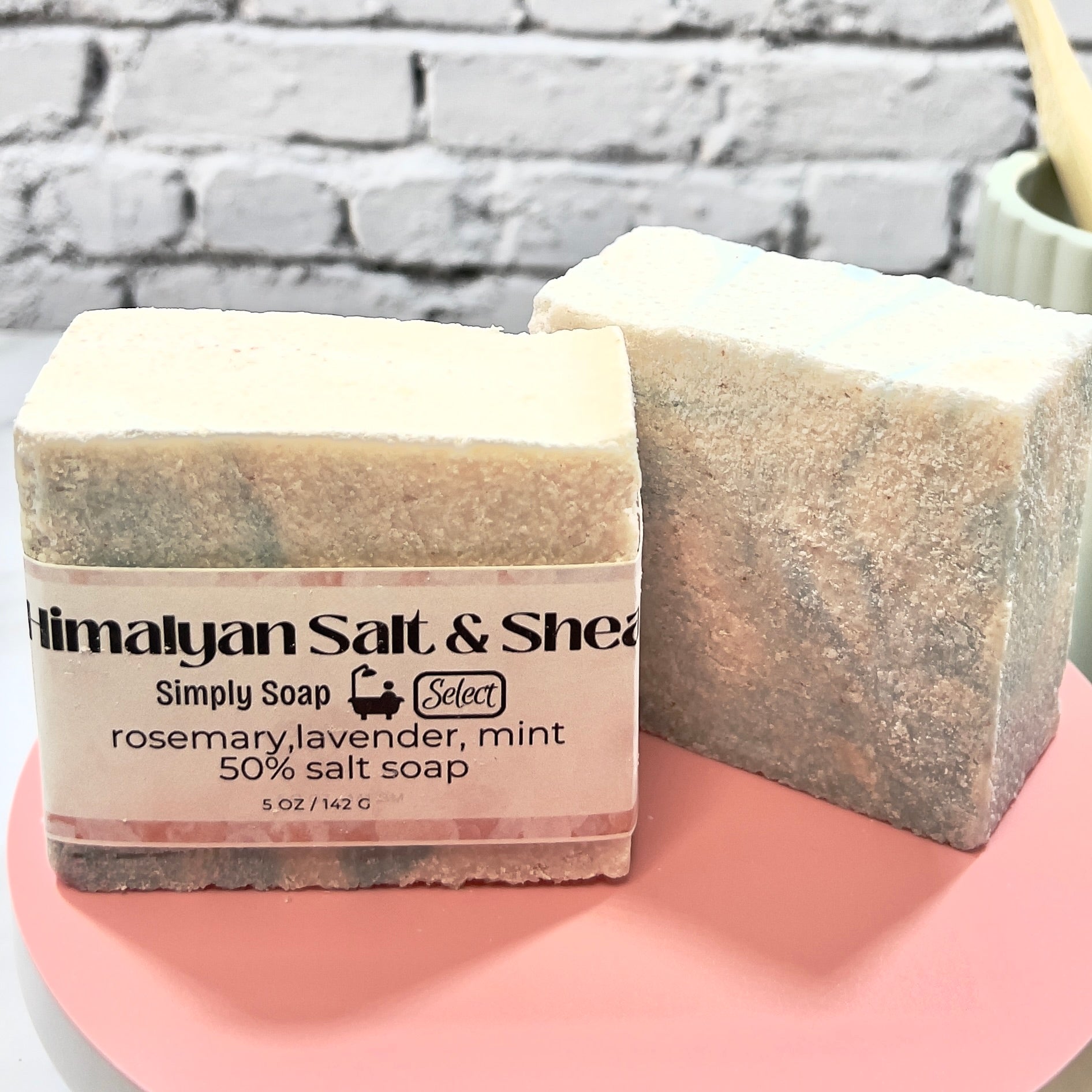 2 Bars of soap with a label on a pink surface against a gray brick wall background