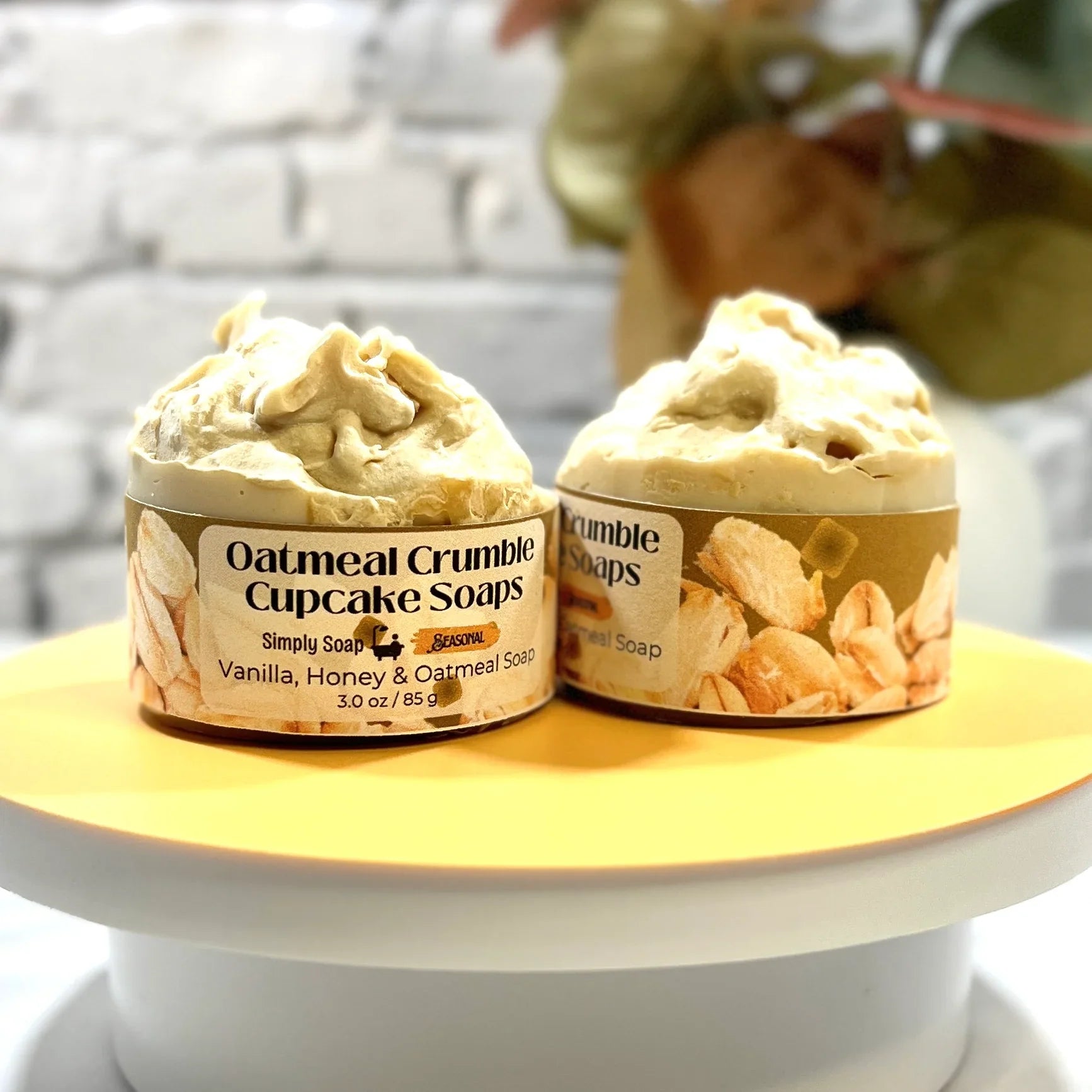 Two Oatmeal Crumble Cupcake Soaps on a yellow stand with a blurred background.