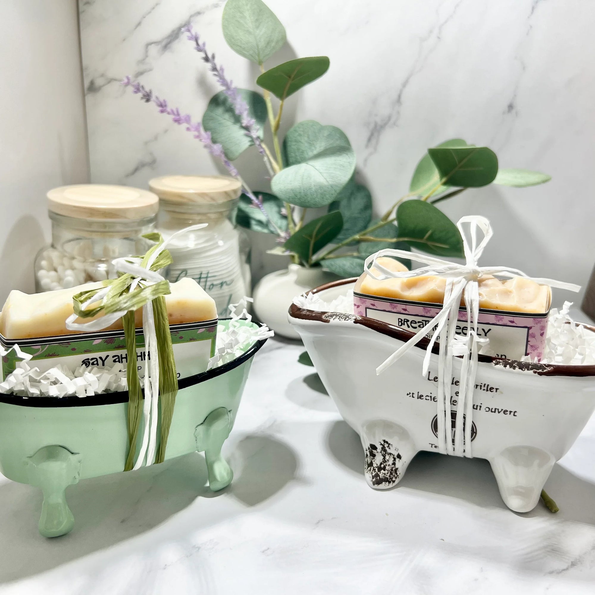 Two decorative bathtub-shaped soap dishes, one in green and one in white, each containing a bar of soap, positioned on a marble surface with a plant and decorative items in the background.
