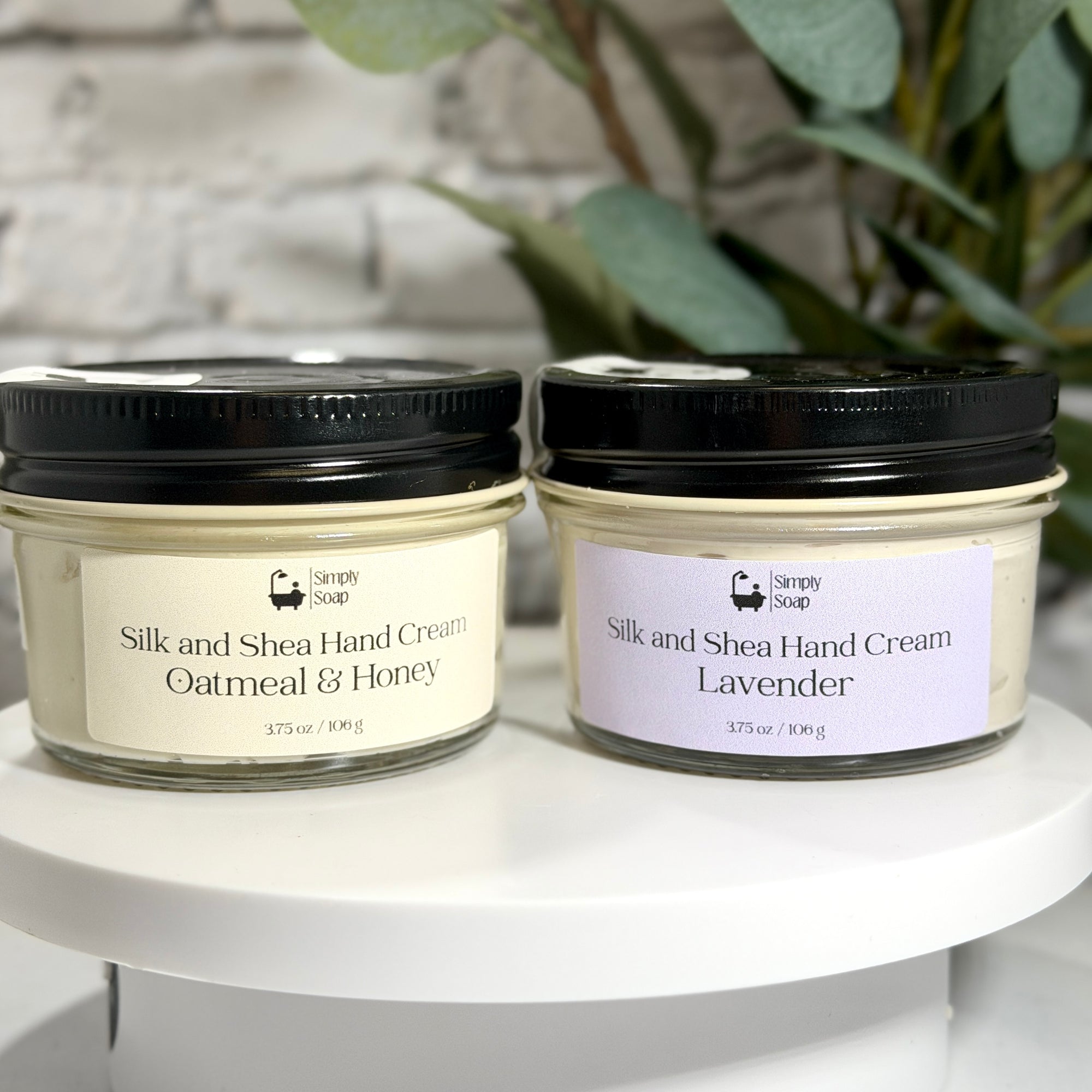 Two jars of Silk and Shea Hand Cream by Simply Soap displayed on a white pedestal. The left jar is labeled 'Oatmeal & Honey' with a cream-colored label and black lid, and the right jar is labeled 'Lavender' with a light lavender label and black lid.