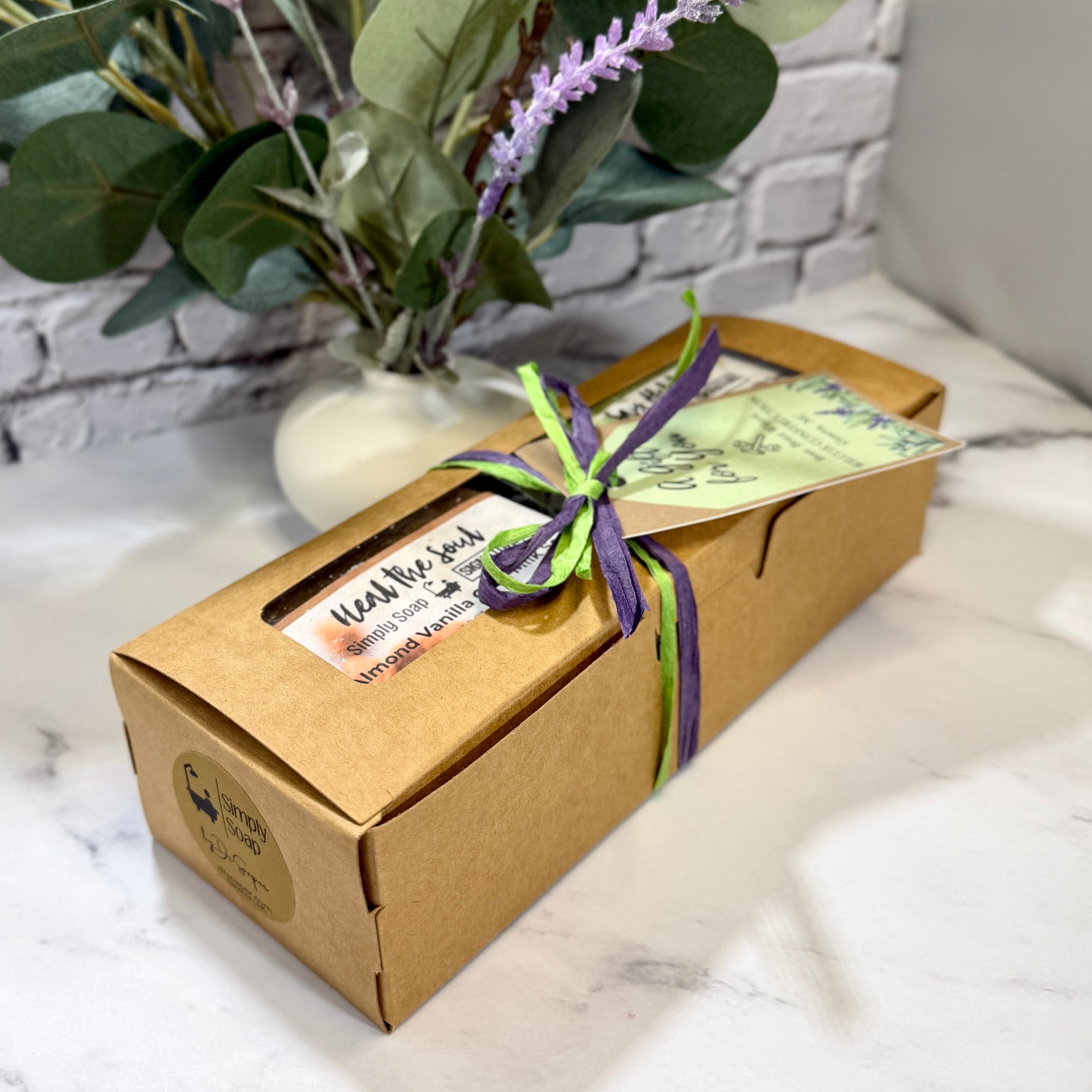 A rectangular kraft paper gift box tied with a green and purple ribbon and a small paper tag, placed on a white marble surface with a white brick wall and a vase of greenery in the background. The box has a round logo sticker on the front left.