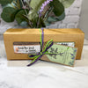 A kraft paper gift box tied with a green and purple ribbon, containing a bar of soap and a decorative tag. Customized with choice of 2 Signature Soaps