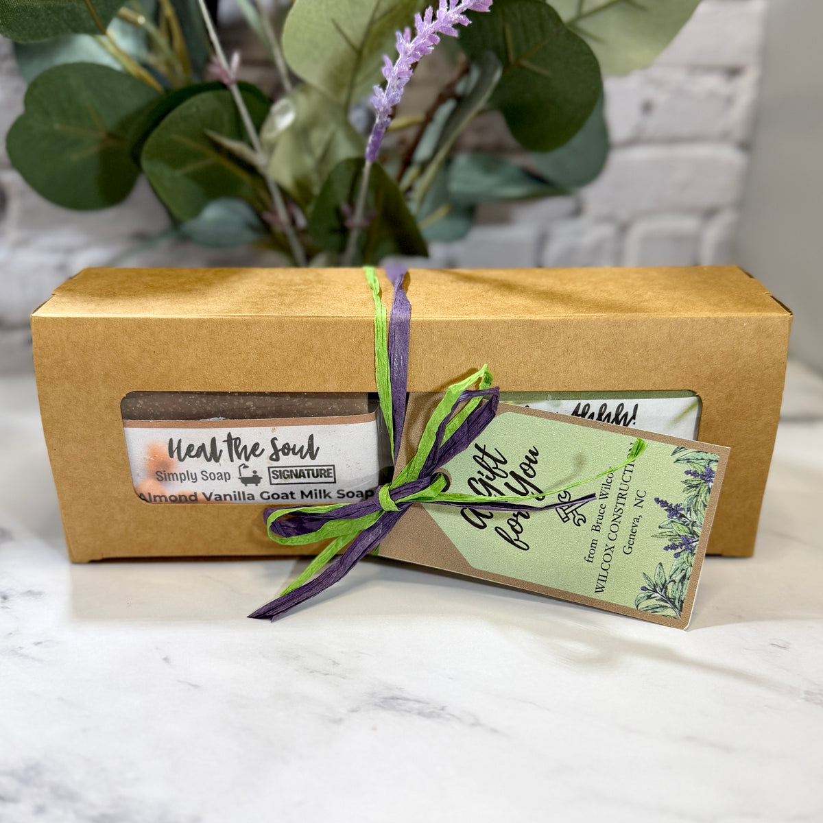 A kraft paper gift box tied with a green and purple ribbon, containing a bar of soap and a decorative tag. Customized with choice of 2 Signature Soaps