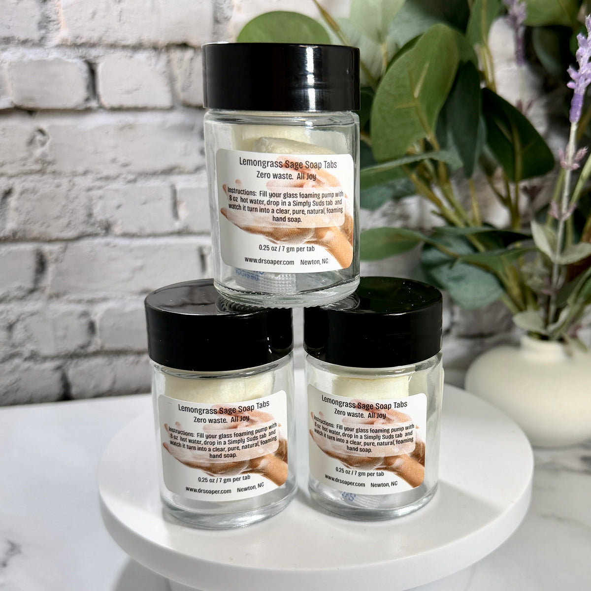 Three glass jars of Lemongrass and Sage Bath Soap Tablets are stacked on a white pedestal. 