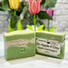 Green soap bar with packaging labeled 'An Apple A Day Apple Champagne' on a white surface with blurred flowers in the background.