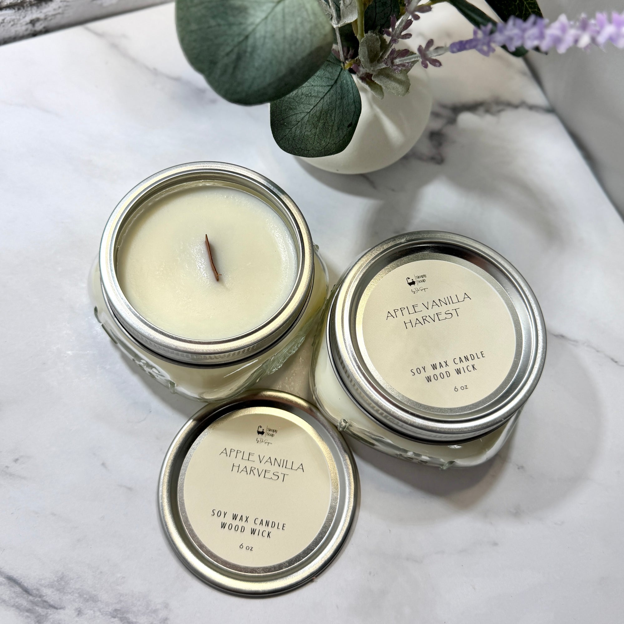 Two 'AppleVanilla Harvest' candles on a marble surface with a plant in the background.