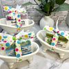 Bars of soap with colorful ribbons in small white bathtubs on a marble surface.