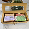 An open kraft gift box containing two holiday bars of soap as a set on a marble surface with a decorative pine cone in the background.