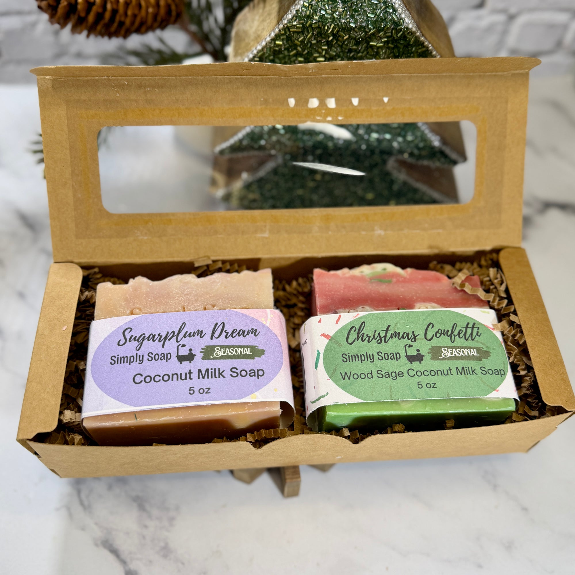 An open kraft gift box containing two holiday bars of soap as a set on a marble surface with a decorative pine cone in the background.