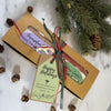 A holiday gift set in a kraft paper box tied with a red and green ribbon and a 'Happy Holidays' tag, displayed on a white marble surface with pine branches and pinecones.