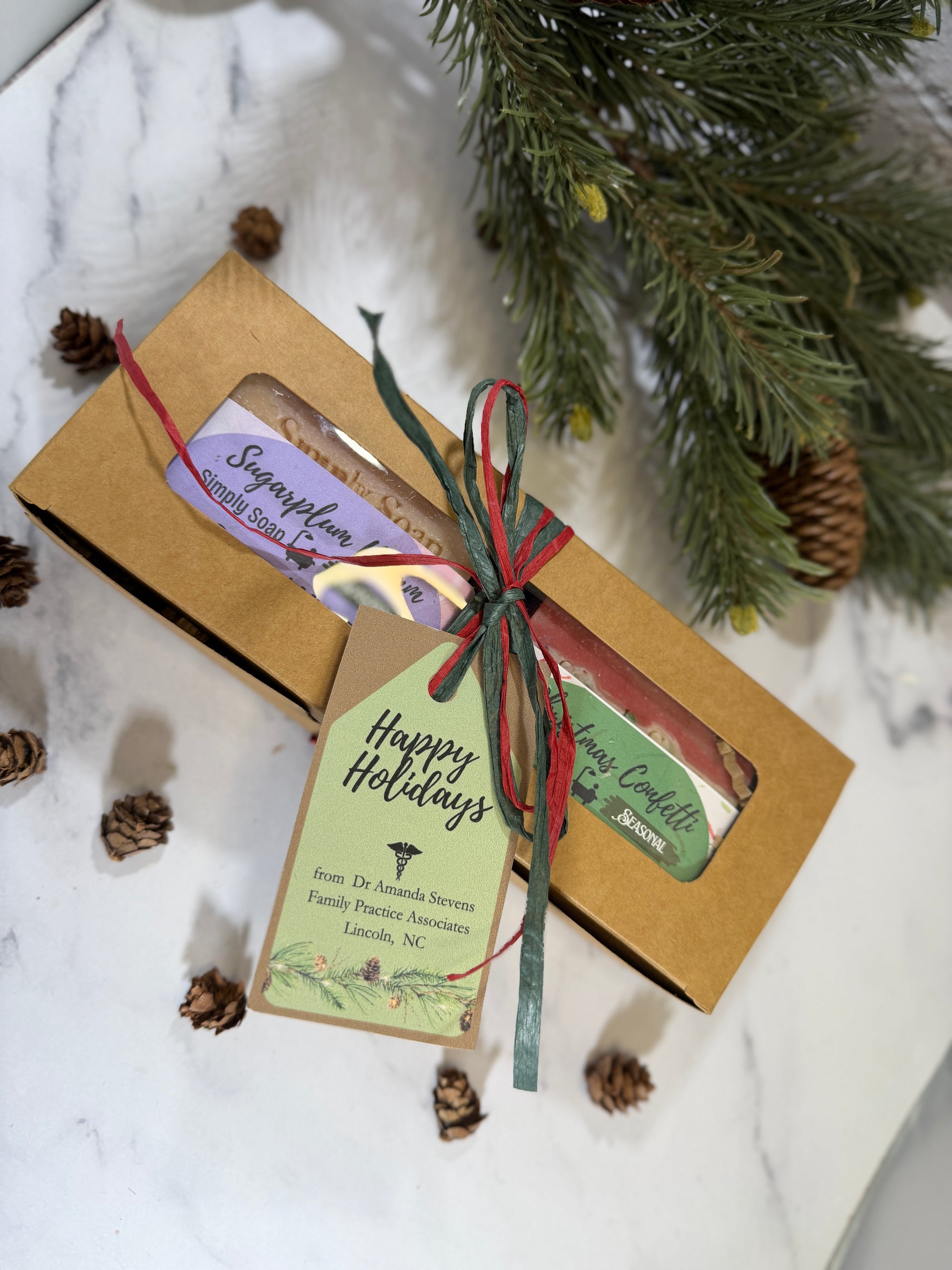 A holiday gift set in a kraft paper box tied with a red and green ribbon and a 'Happy Holidays' tag, displayed on a white marble surface with pine branches and pinecones.