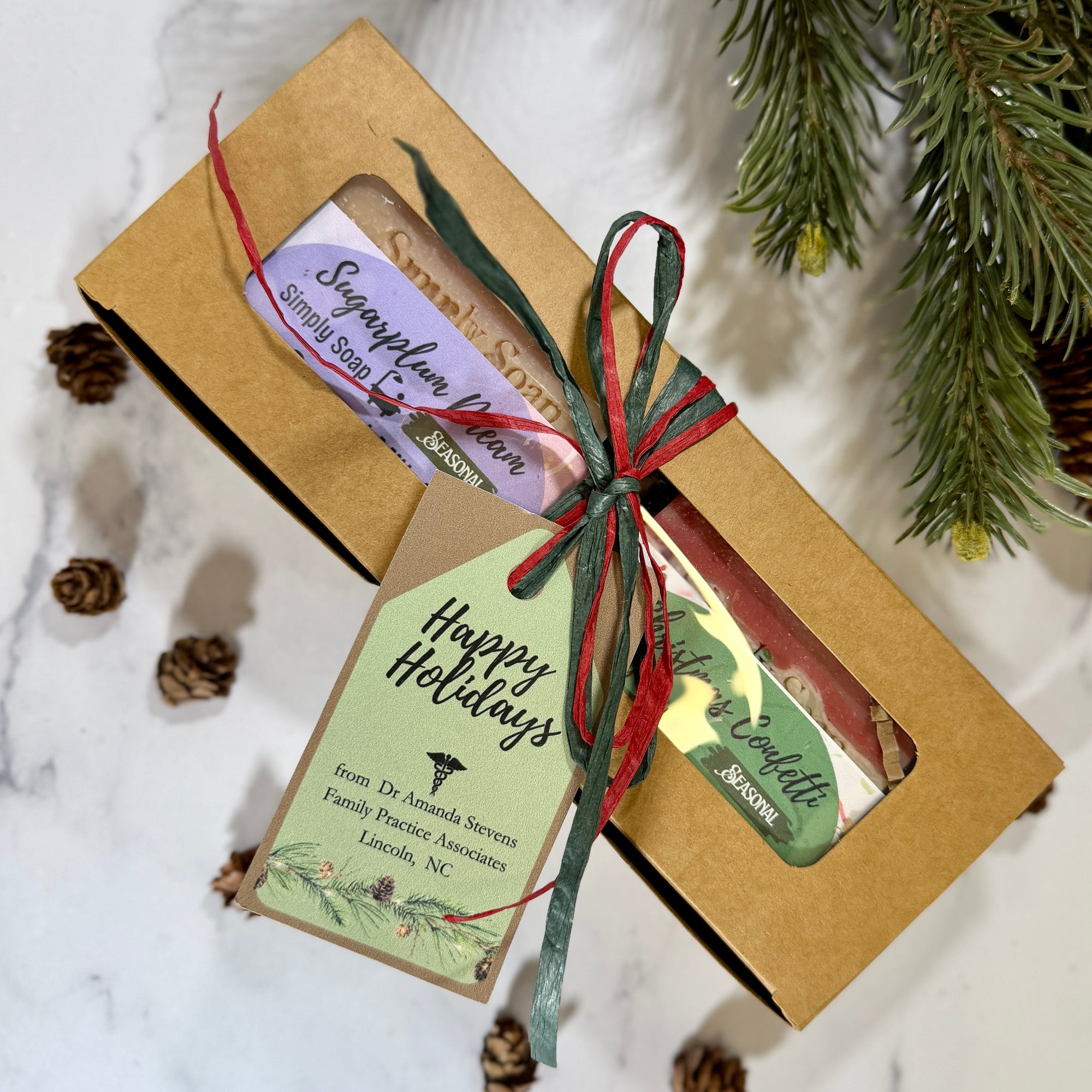 A gift set of 2 holiday handmade soaps in a kraft paper box closes and  tied with a red and green ribbon. 