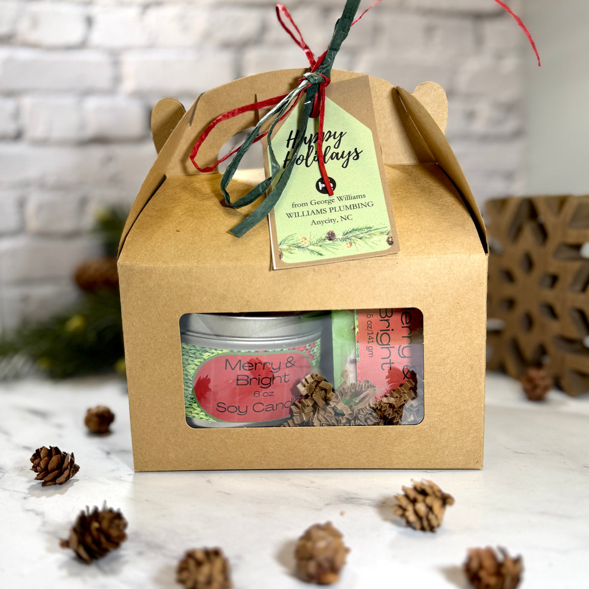 A kraft paper gift box with a clear front window displaying soap and  soy candle  with ribbon and gift tag in gable box. 