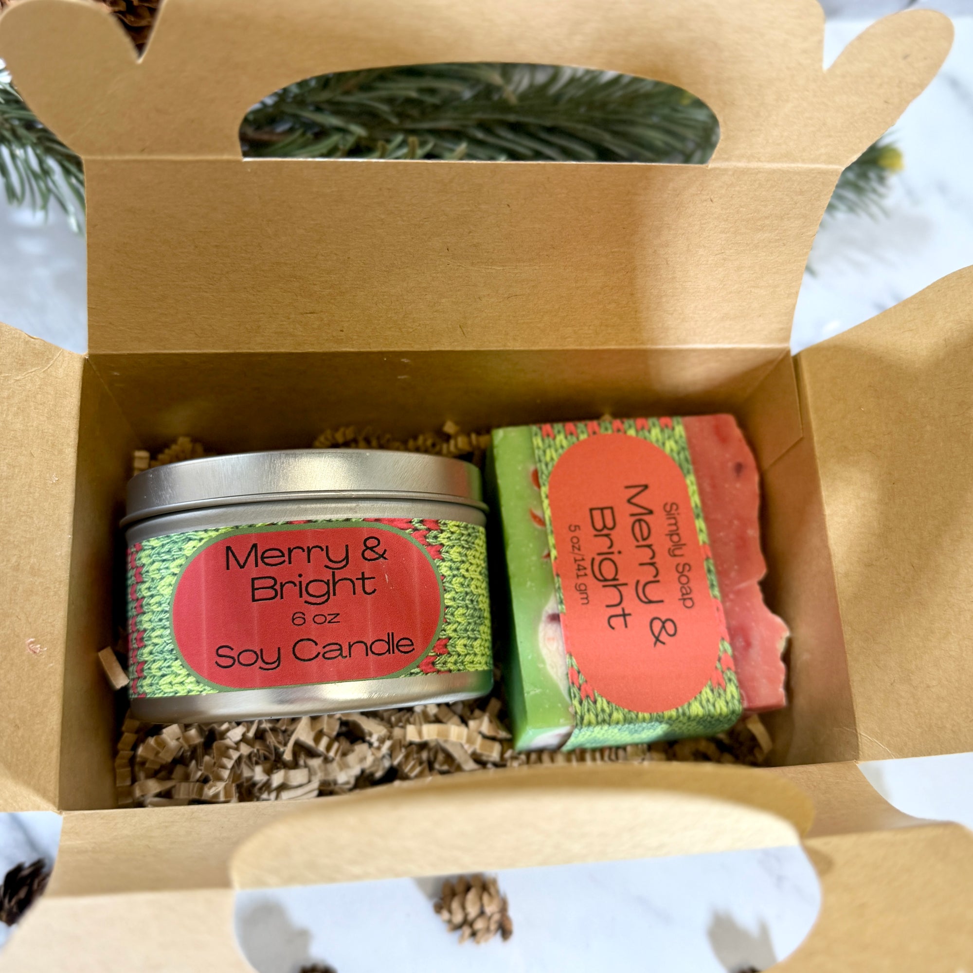 A gift set in an open kraft gable paper box containing a silver tin candle and a  bar soap, for Christmas.