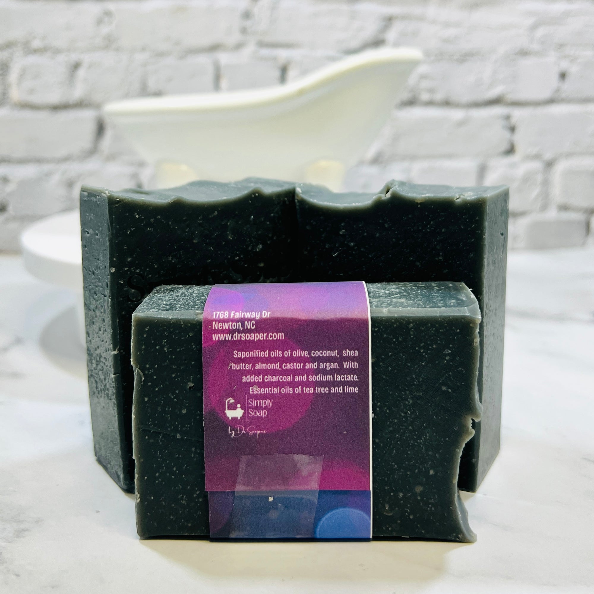 Black soap bar with a purple label on a white surface with a gray brick wall background