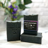 Black soap bars with 'Simply Soap' branding on a white surface with blurred greenery in the background.