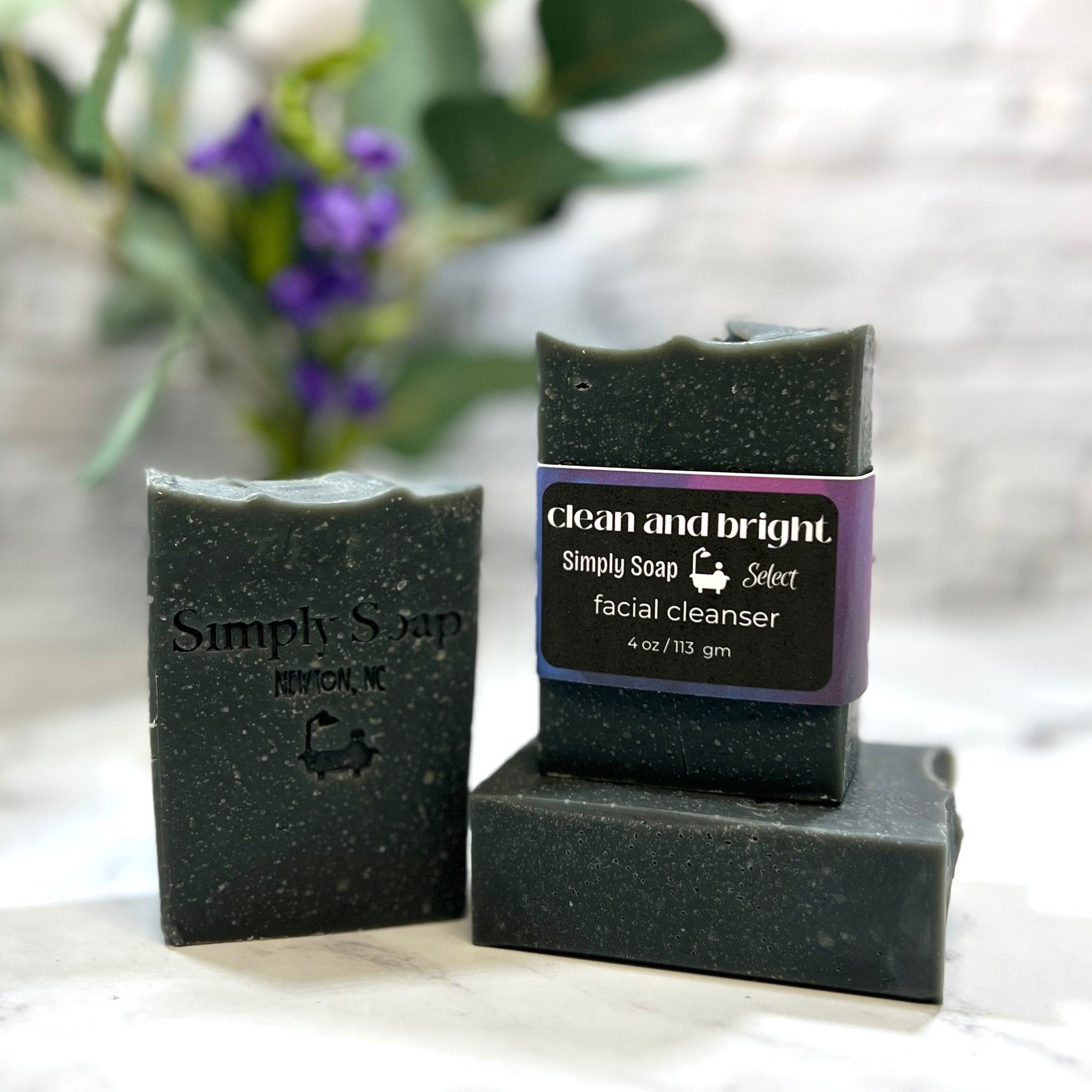 Black soap bars with 'Simply Soap' branding on a white surface with blurred greenery in the background.
