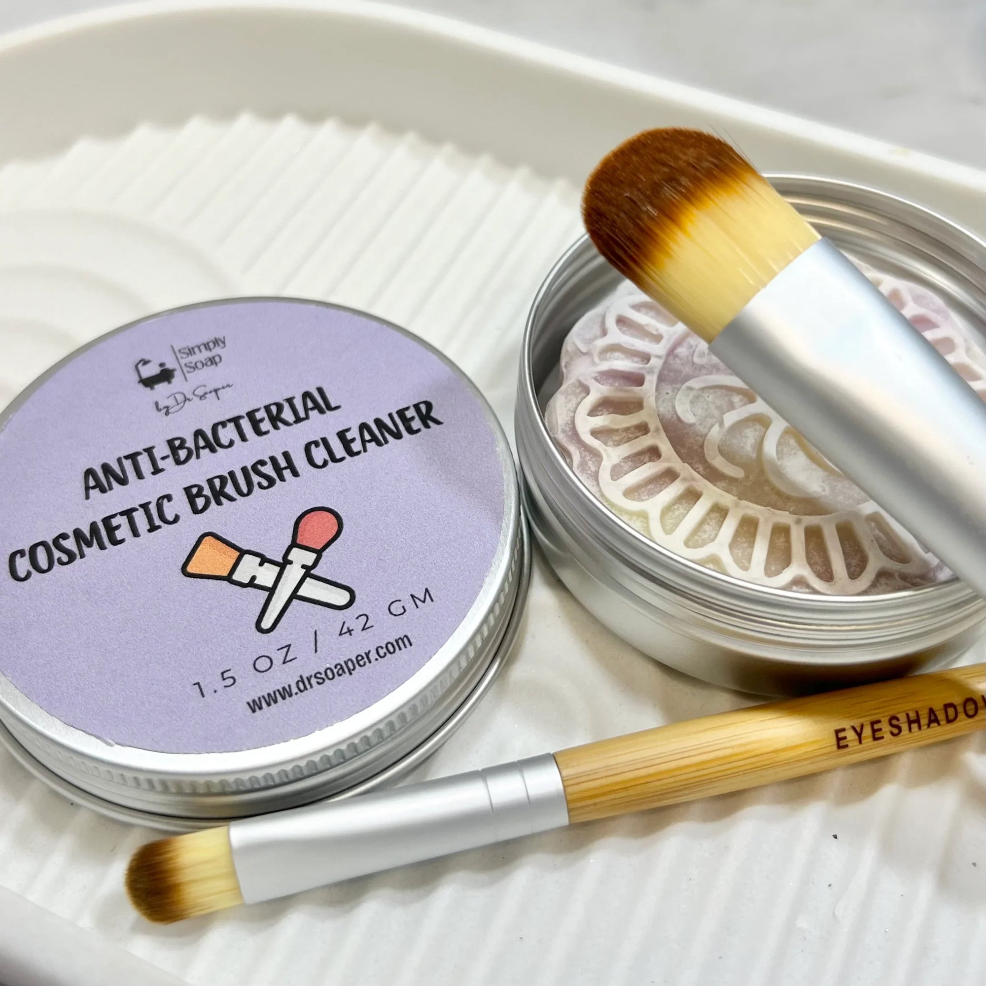 Dr Soaper's Cosmetic Brush Cleaner open with lid next to tin and brush on tip