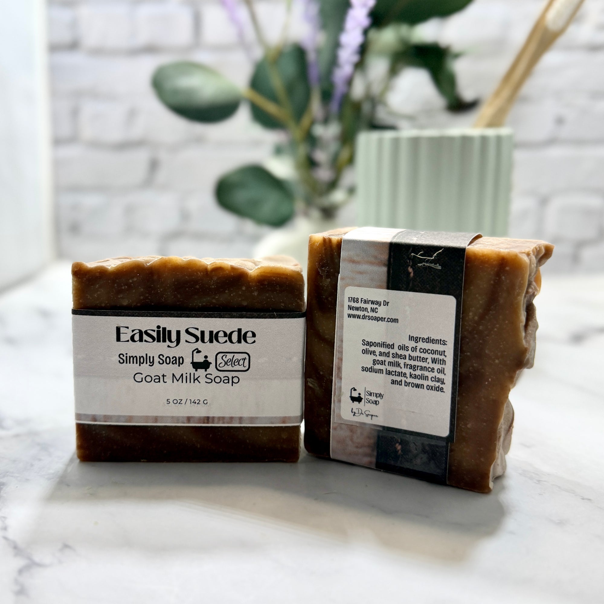 Two bars of goat milk soap with labels on a marble surface, with a plant in the background.
