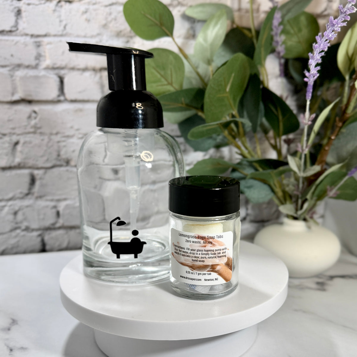 A clear glass bath foam bottle with a black pump and a small clear jar with a black lid filled with 3 soap tabs. 