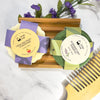 Two soap bars with decorative labels on a wooden tray, surrounded by flowers and a comb on a marble surface.