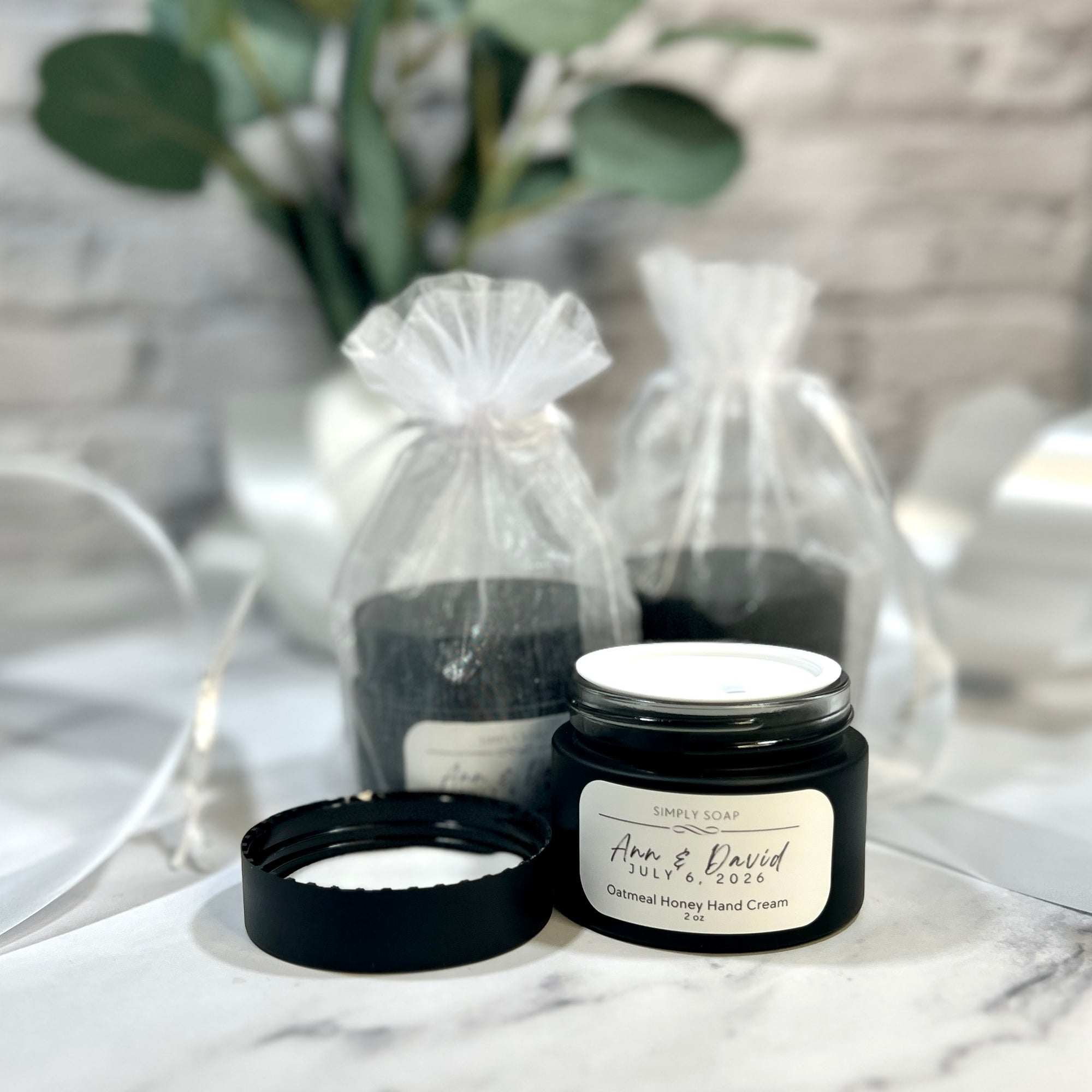Hand cream jar with a label on a marble surface, with gift bags and plants in the background.