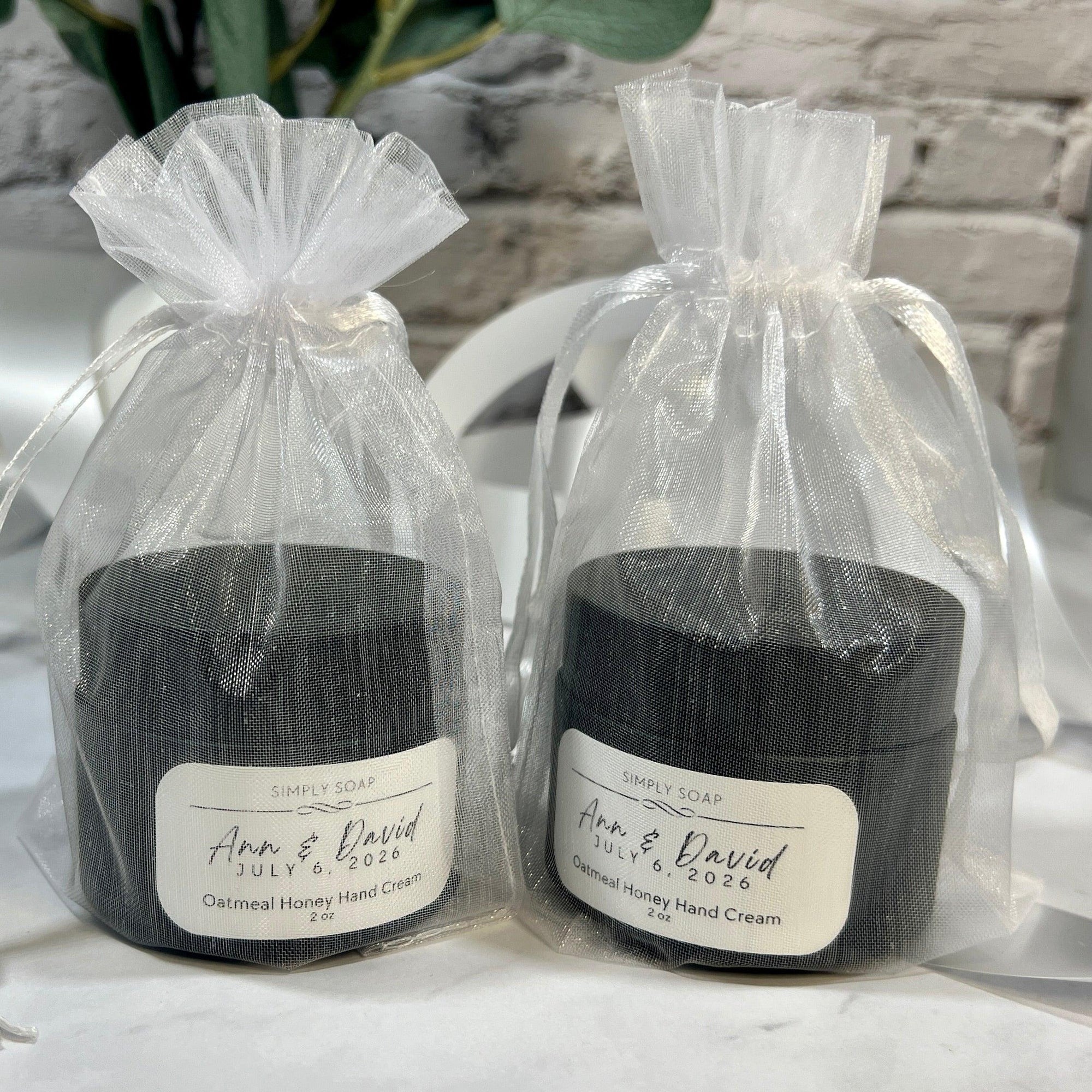 Two black containers of hand cream in clear gift bags with labels on a marble surface.