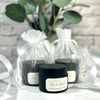 Black jars of cream with white labels on a marble surface, surrounded by sheer white ribbons and greenery.