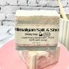 Bar of soap with a label on a pink stand against a gray brick wall.