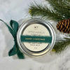 Candle jar labeled 'Apple Vanilla Harvest' with a green ribbon and pine branches on a white background
