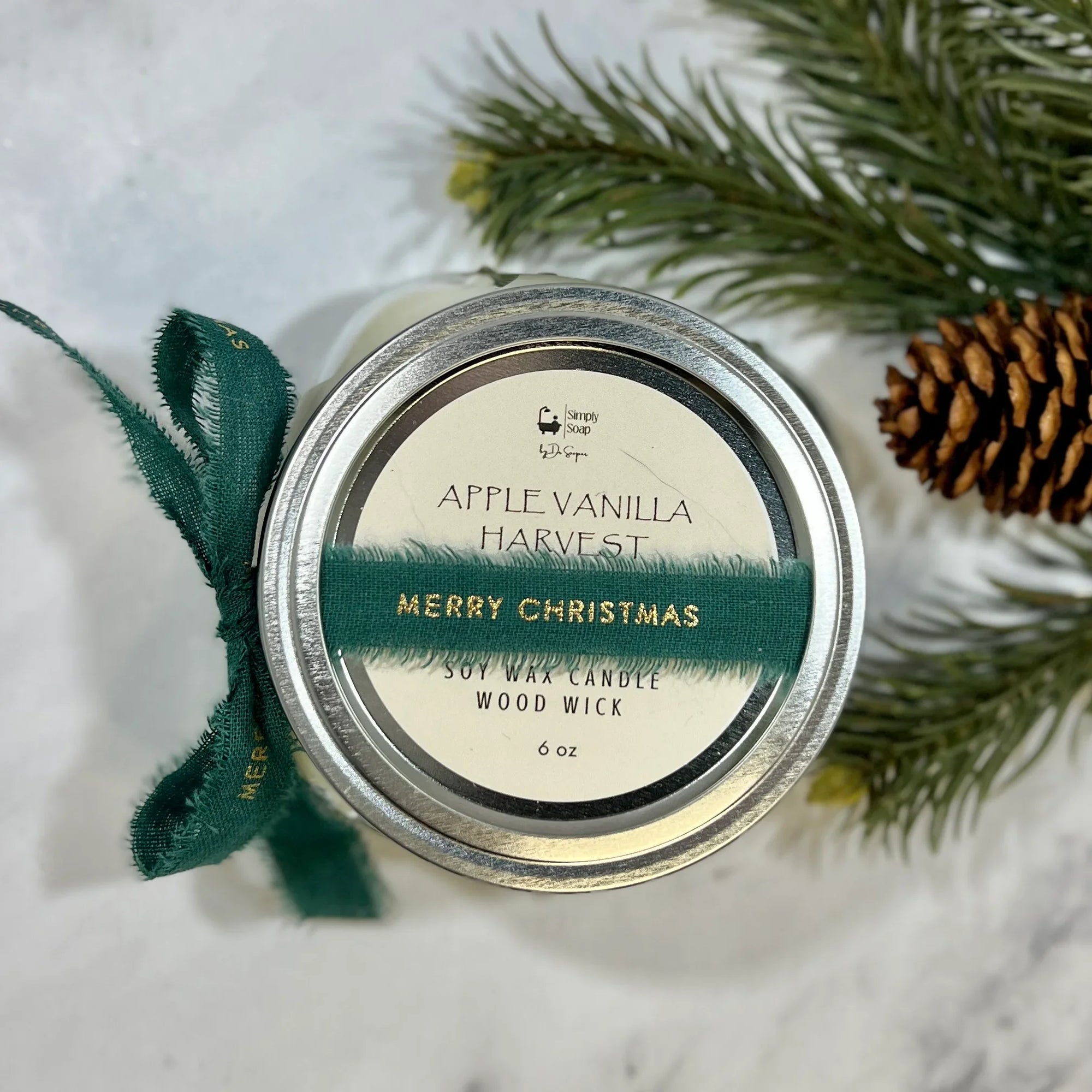 Candle jar labeled 'Apple Vanilla Harvest' with a green ribbon and pine branches on a white background
