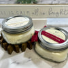 Two jars of candles with labels on a marble surface, with a decorative sign in the background.