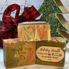 Three bars of goat milk soap with 'Simply Soap' and 'Holiday Hearth' labels, Christmas tree and plaid fabric in the background.