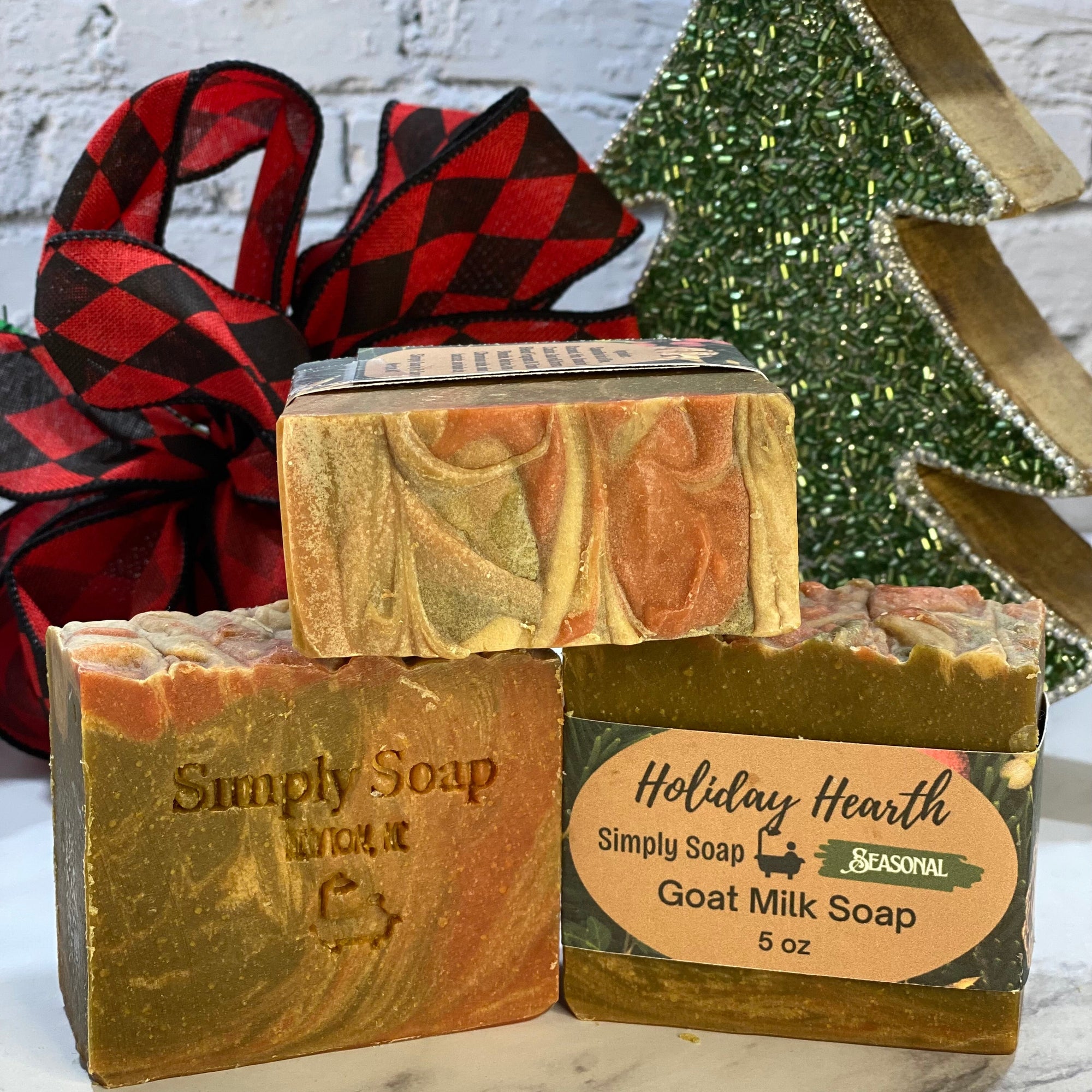 Three bars of goat milk soap with 'Simply Soap' and 'Holiday Hearth' labels, Christmas tree and plaid fabric in the background.