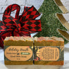 Two bars of goat milk soap with holiday-themed packaging in front of a decorative Christmas tree and plaid fabric.