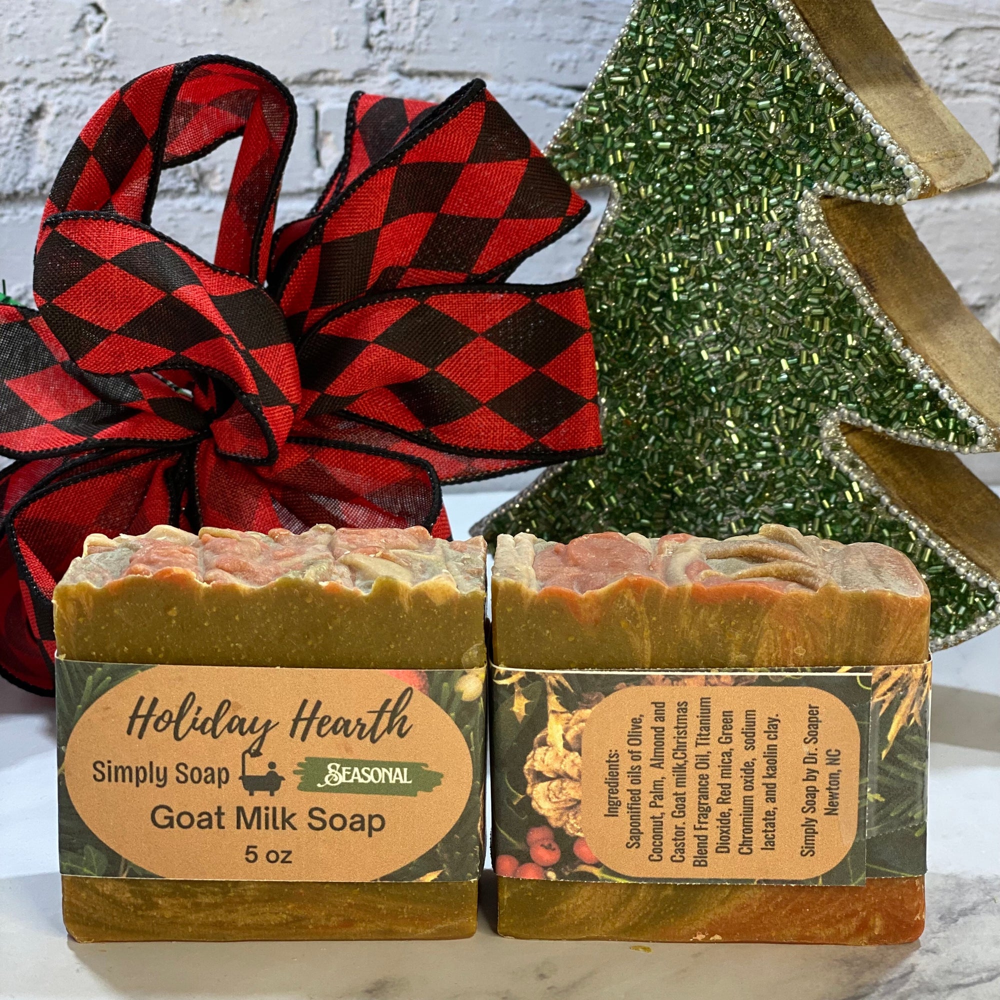 Two bars of goat milk soap with holiday-themed packaging in front of a decorative Christmas tree and plaid fabric.
