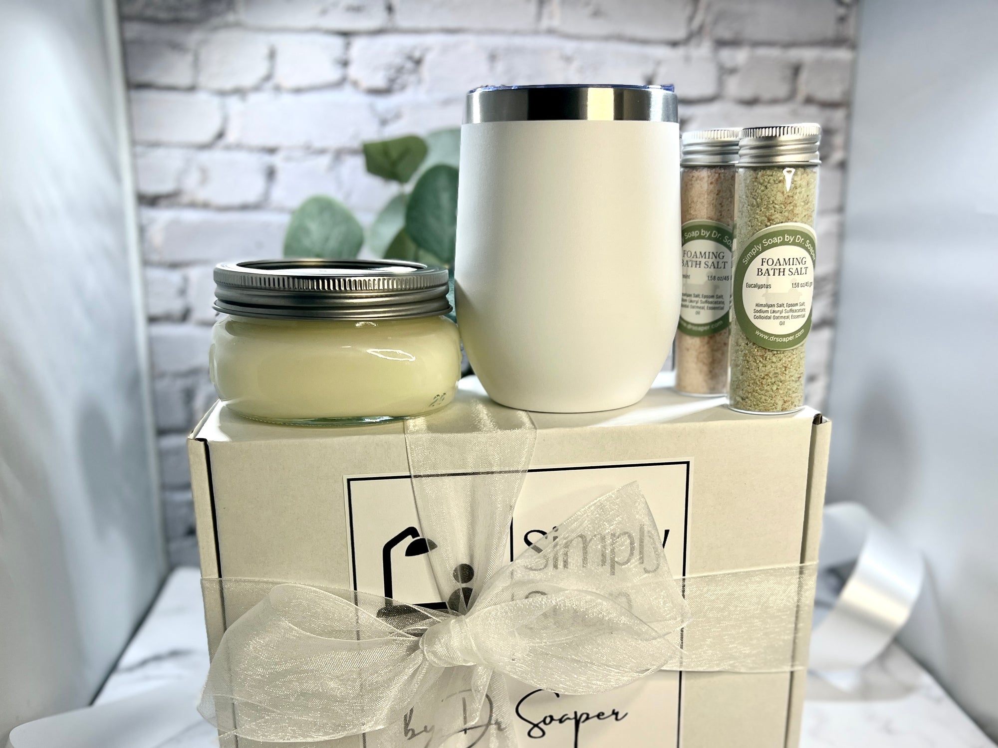 Gift set with a candle, tumbler, and bath salts on a white surface with a blurred background.