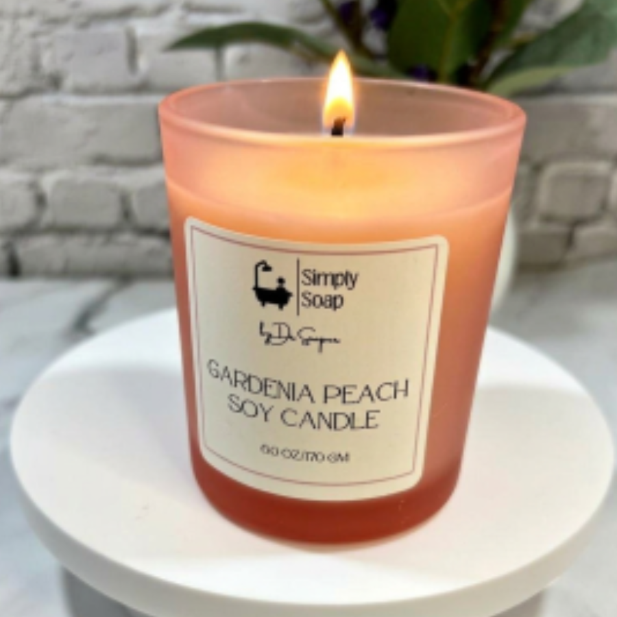 A lit pink colored soy candle in a frosted glass jar with a white label reading 'Simply Soap by Dr. Soaper Gardenia Peach Soy Candle,' placed on a white round pedestal against a blurred white brick wall and green plant background.