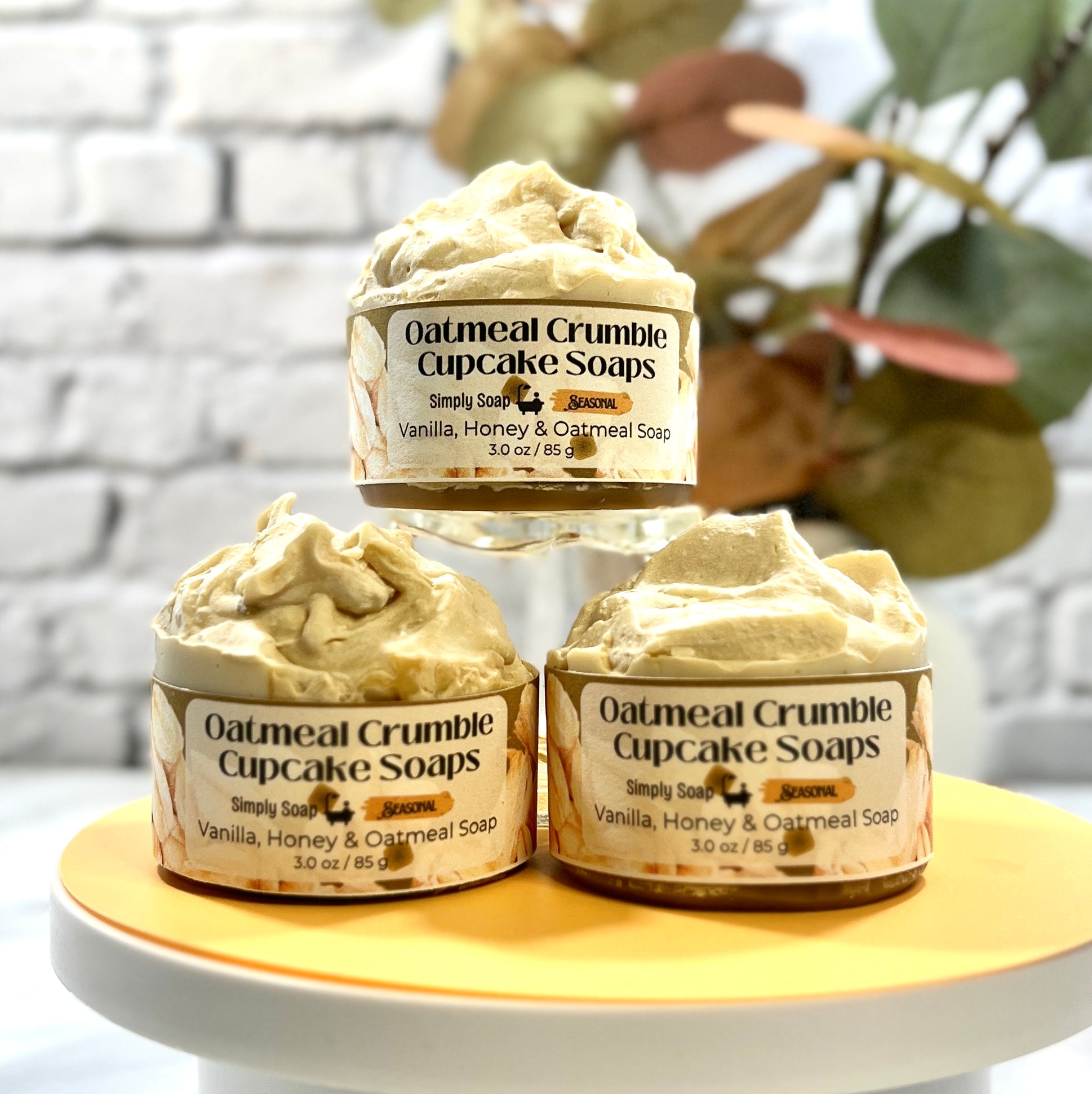 Three Oatmeal Crumble Cupcake Soaps on a white stand with a blurred plant background
