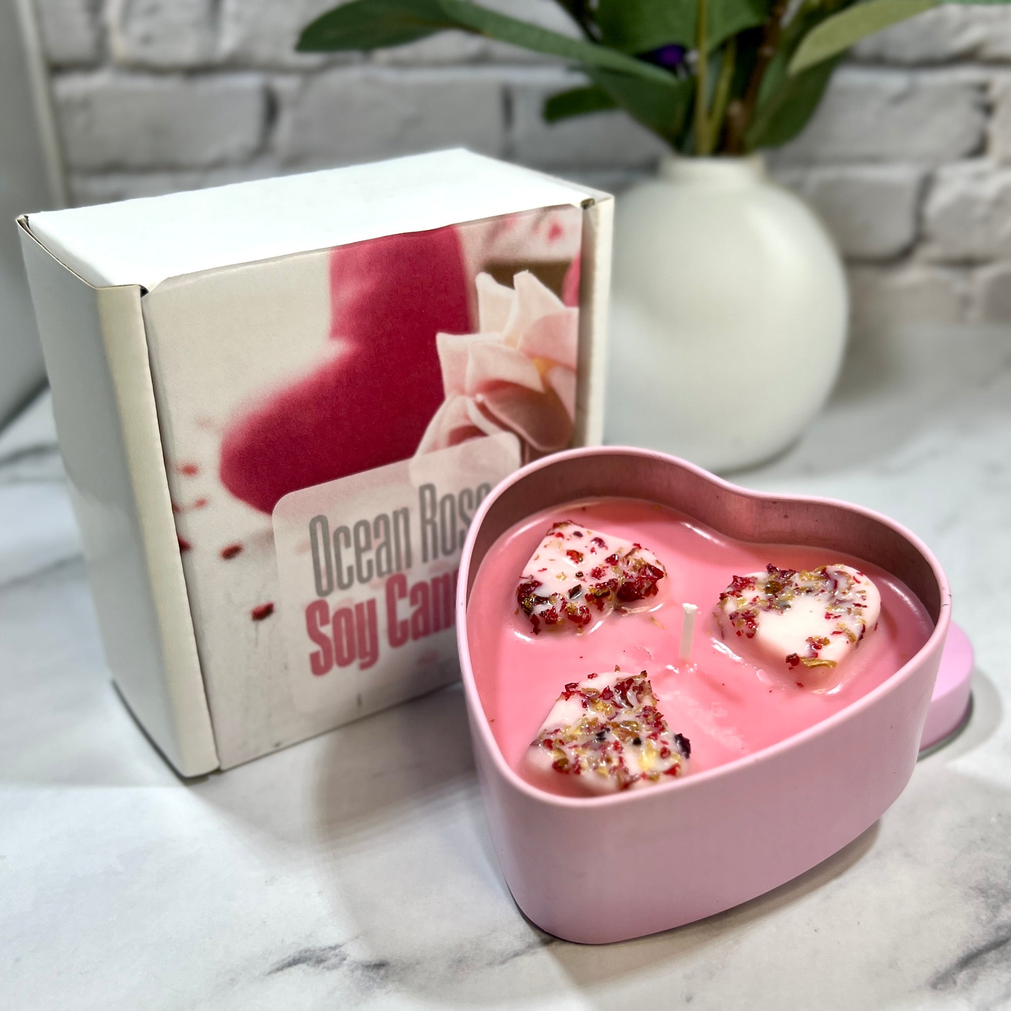 Heart-shaped pink container with ocean rose soy candles next to a branded box on a marble surface.