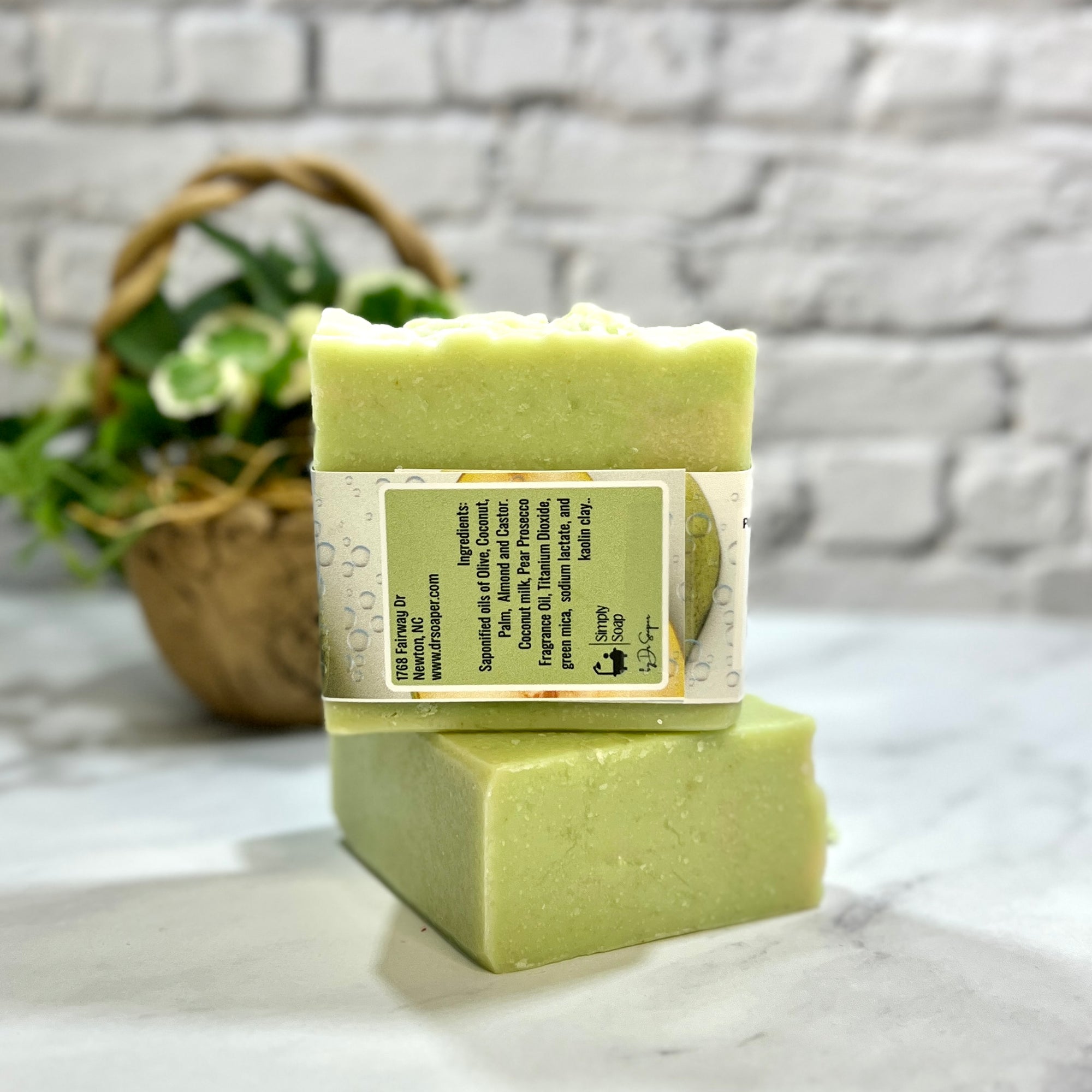 Two bars of green soap stacked on a marble surface with a basket and plant in the background.