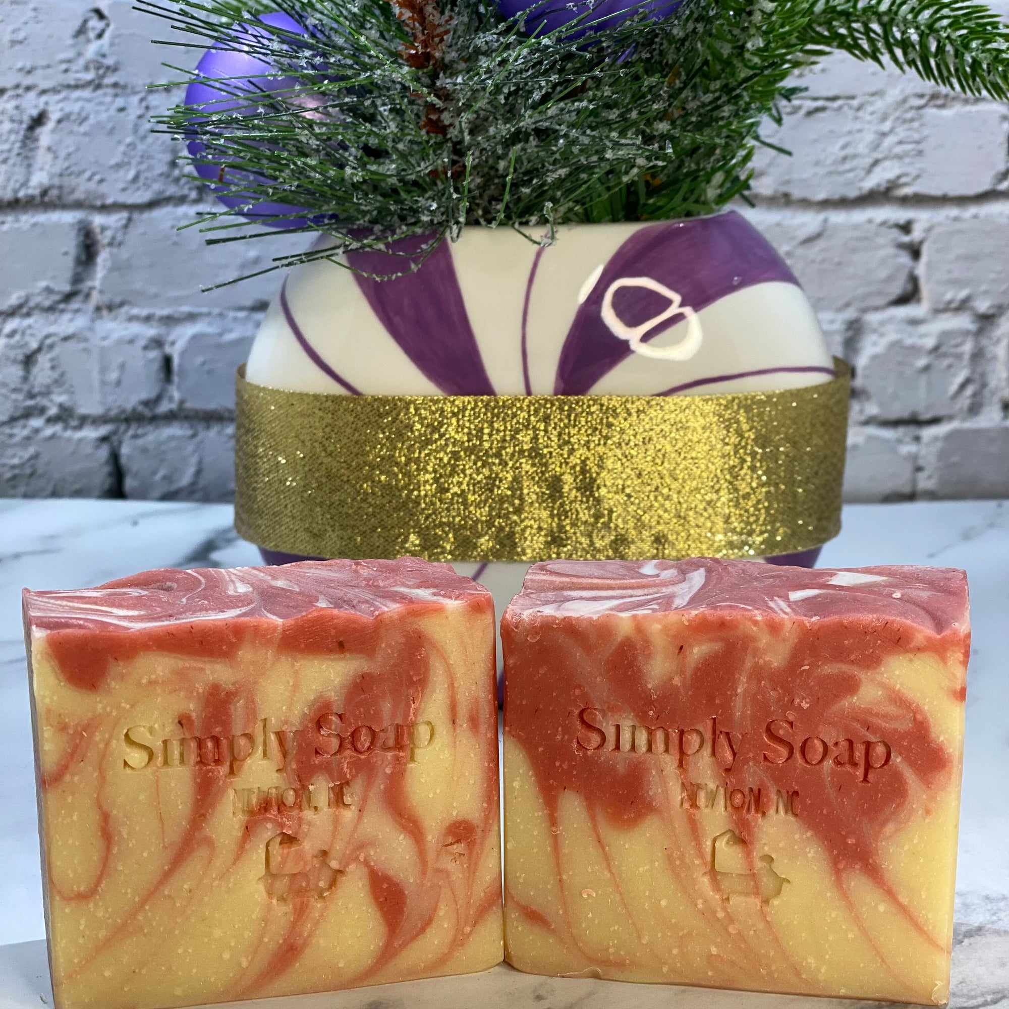 Two red marbled soap bars labeled 'Simply Soap' with a decorative pot and ornaments in the background.