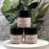 Three small jars of Powder & Petals Face Wash by The Beauty Shop are arranged in a pyramid on a white round tray, with blurred green leaves and purple flowers in the background.