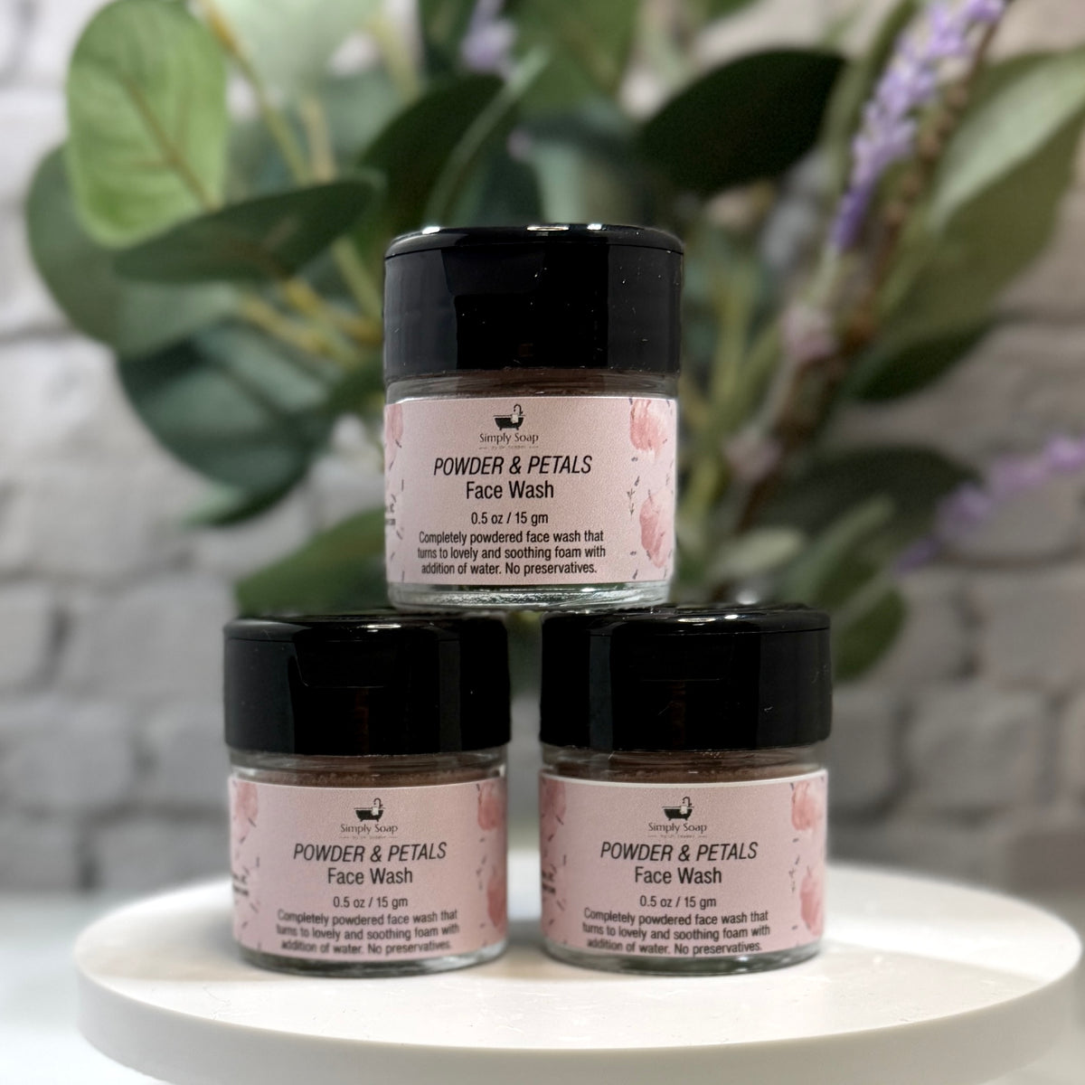 Three small jars of Powder & Petals Face Wash by The Beauty Shop are arranged in a pyramid on a white round tray, with blurred green leaves and purple flowers in the background.