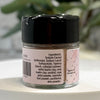 A small clear jar of face powder with a black lid and a pink label, shown on a white surface with a blurred green plant in the background. The label lists ingredients .