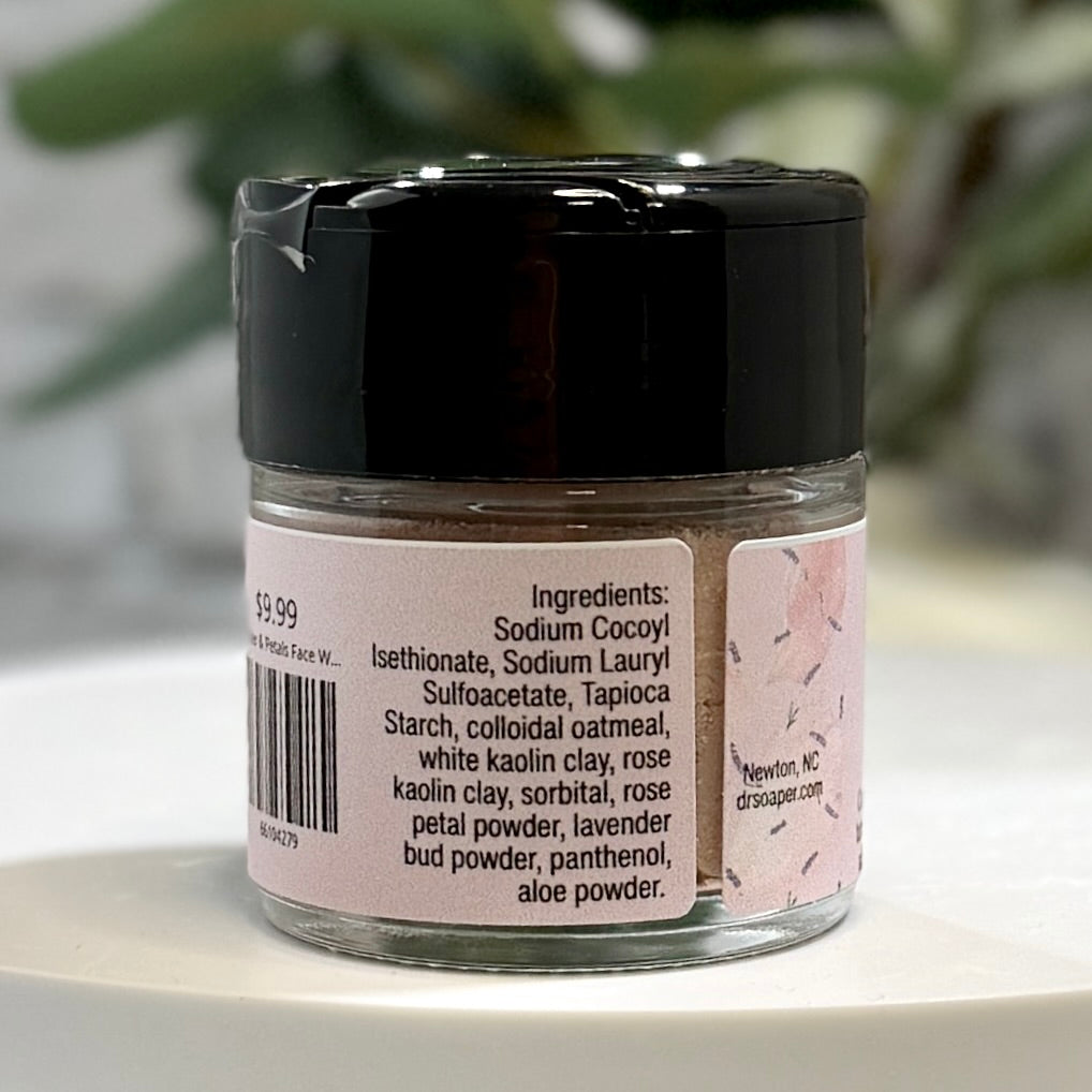 A small clear jar of face powder with a black lid and a pink label, shown on a white surface with a blurred green plant in the background. The label lists ingredients .