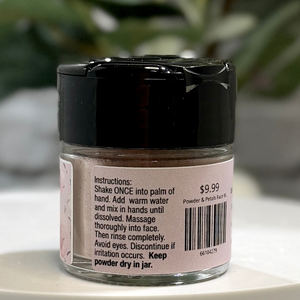 A small clear jar of facial powder with a black lid and a pink label showing product instructions and a barcode, photographed on a light surface with a blurred green plant background.