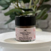 A small clear jar of face wash with a black lid and a pink label reading 'Powder & Petals Face Wash,' displayed on a white round surface with blurred green foliage in the background.