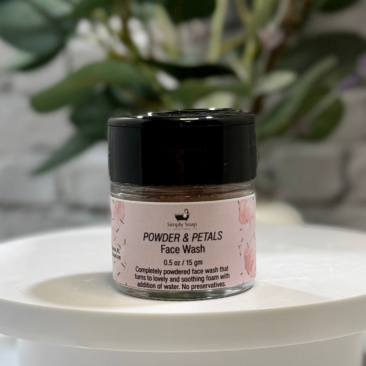 A small clear jar of face wash with a black lid and a pink label reading 'Powder & Petals Face Wash,' displayed on a white round surface with blurred green foliage in the background.