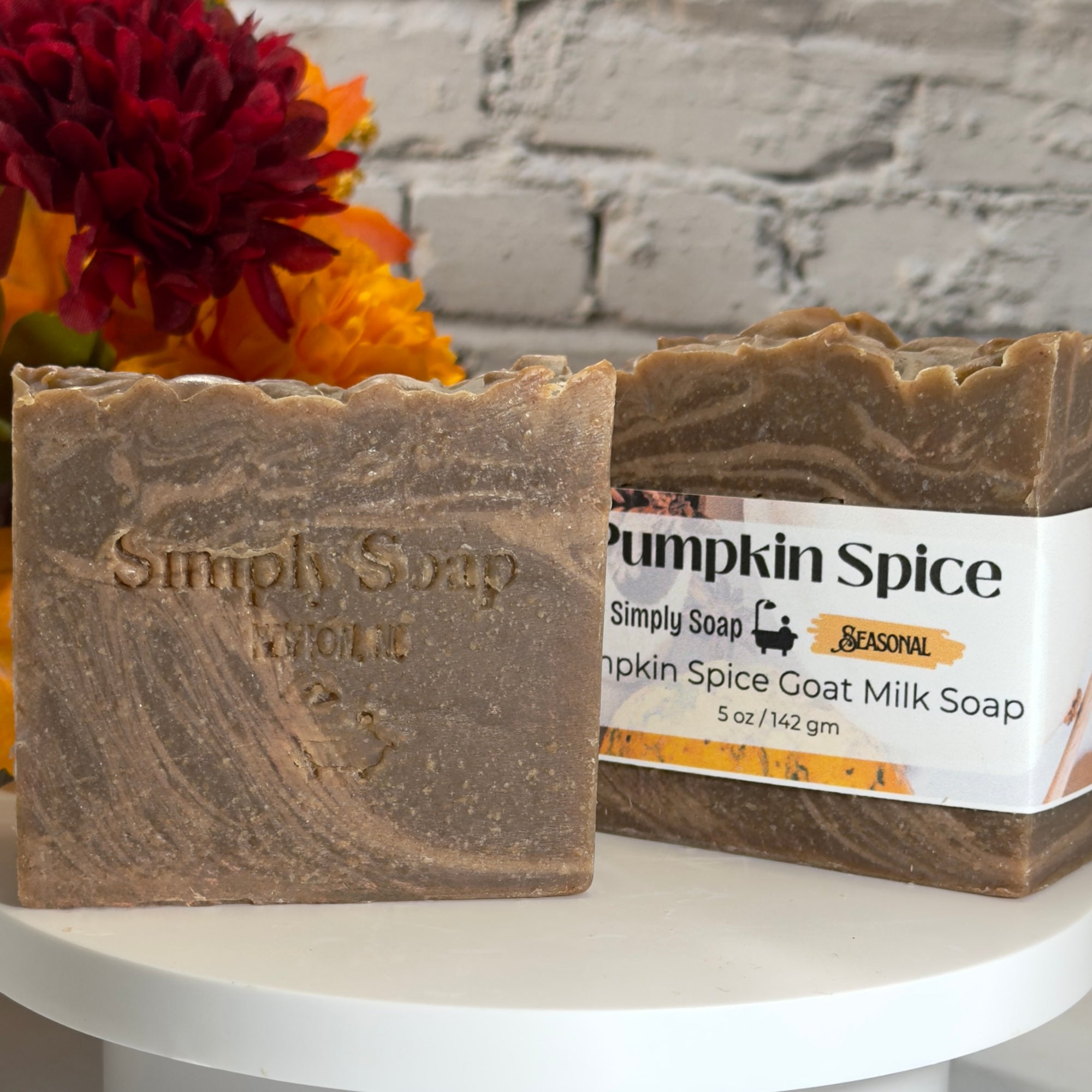Two bars of Simply Soap 'Pumpkin Spice' goat milk soap displayed on a white round table, with a rustic white brick wall and orange and red flowers in the background.